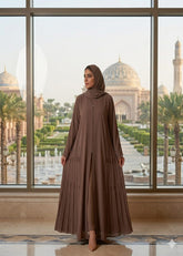 Elegant woman wearing a premium pleated taupe abaya set with matching hijab standing in front of a scenic architectural window.