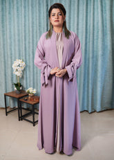 Woman wearing a light purple abaya standing in front of a blue curtain with a small table and flowers in the background.