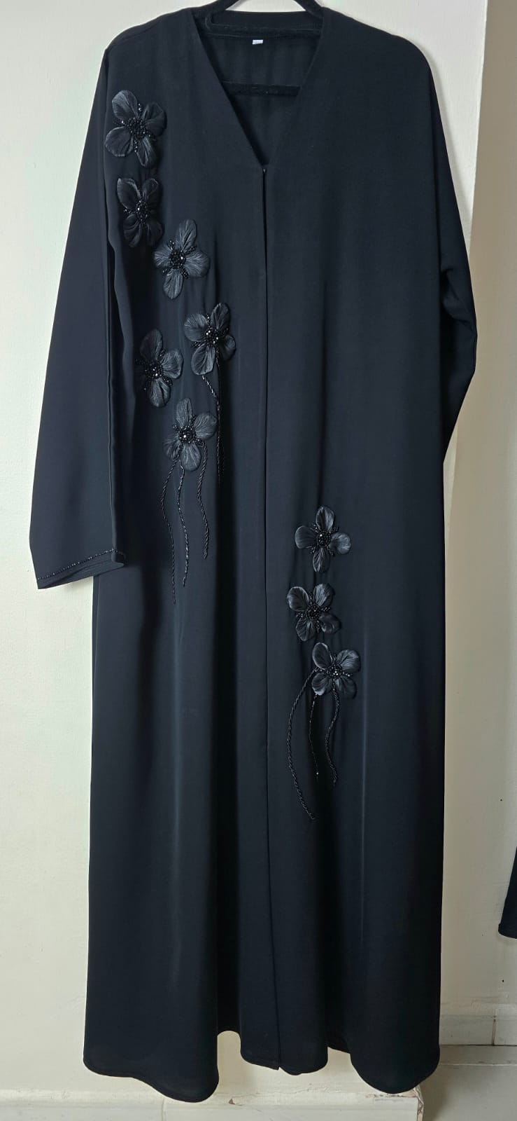 A long black abaya dress with floral embroidery on the front, hanging against a plain off-white wall.