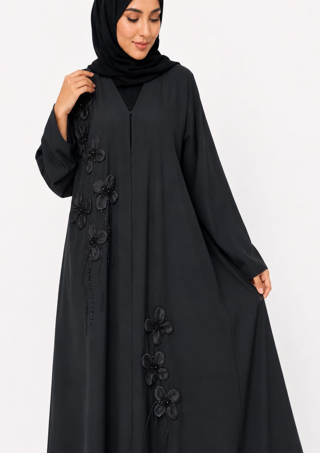 A woman wearing a long black abaya with 3D floral appliqué details and a matching black hijab, photographed against a plain white studio background.