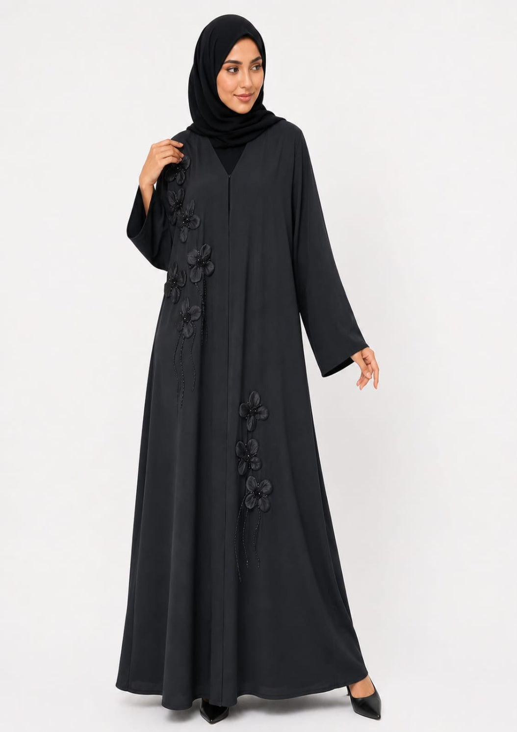 A woman wearing a long black abaya with 3D floral applique and a matching black hijab, standing against a plain light gray studio background.