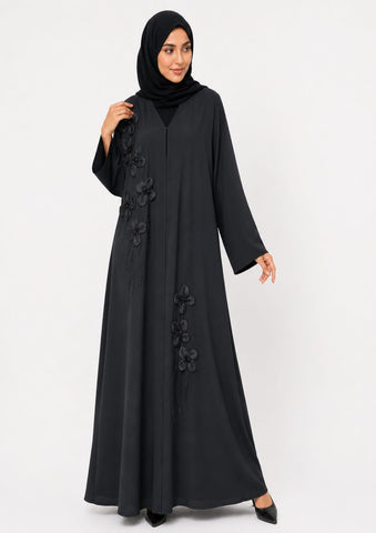 A woman wearing a long black abaya with 3D floral applique and a matching black hijab, standing against a plain light gray studio background.
