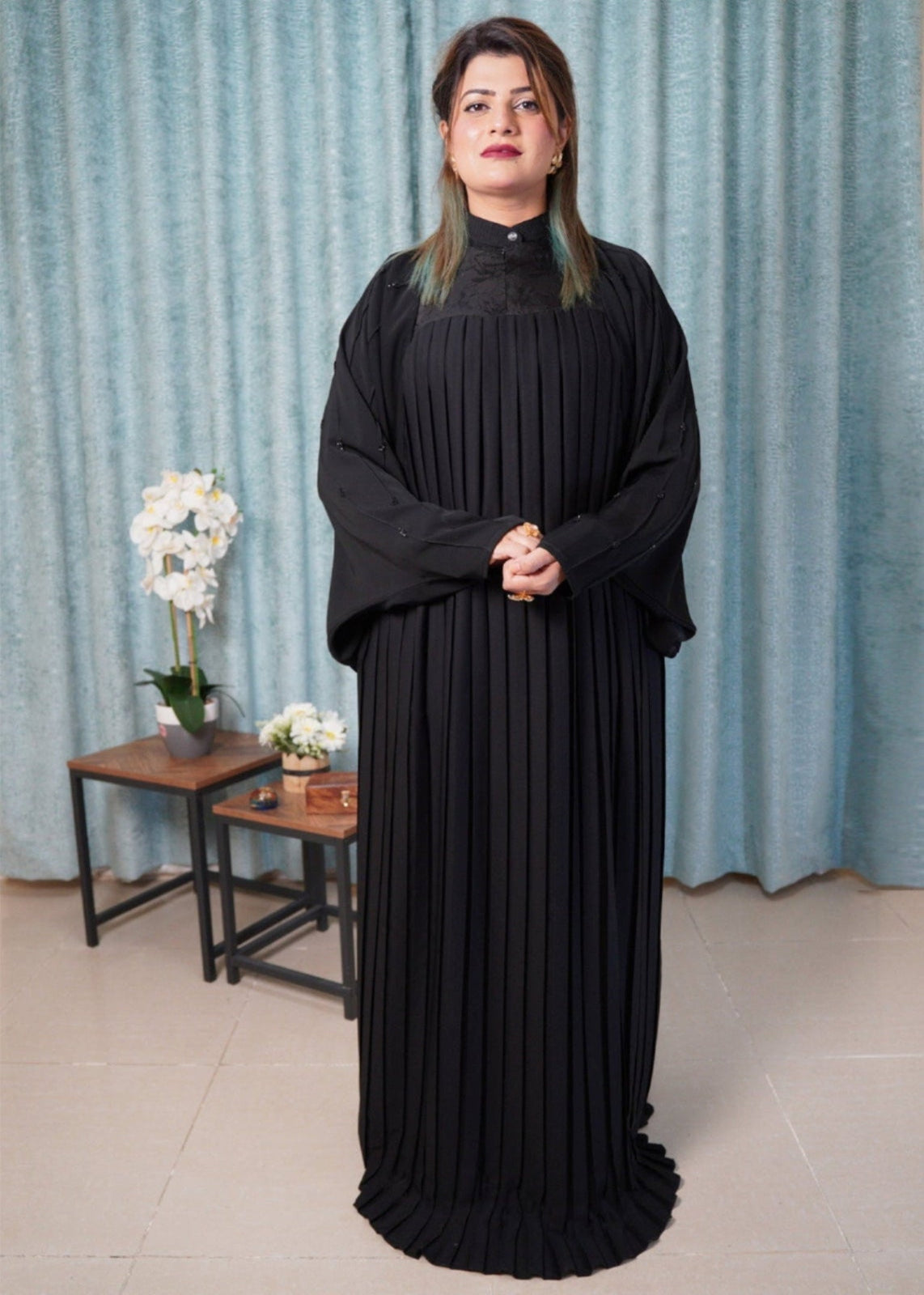Woman wearing a black dress standing in a room with light blue curtains and a small table with flowers.