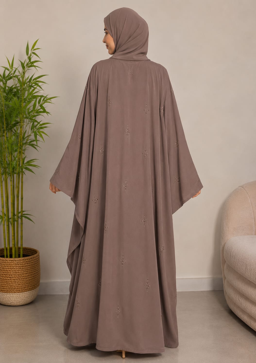 A woman wearing a taupe brown modest dress with a matching hijab, shown from the back in a minimalist indoor setting with a potted plant and a light-colored sofa.