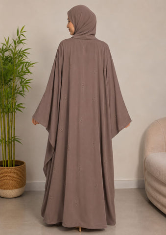A woman wearing a taupe brown modest dress with a matching hijab, shown from the back in a minimalist indoor setting with a potted plant and a light-colored sofa.